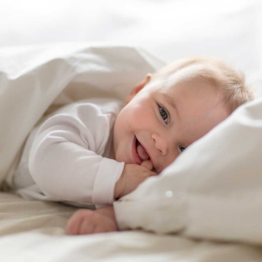 Lächelndes Baby liegt unter einer Decke und greift an die eigenen Hände.